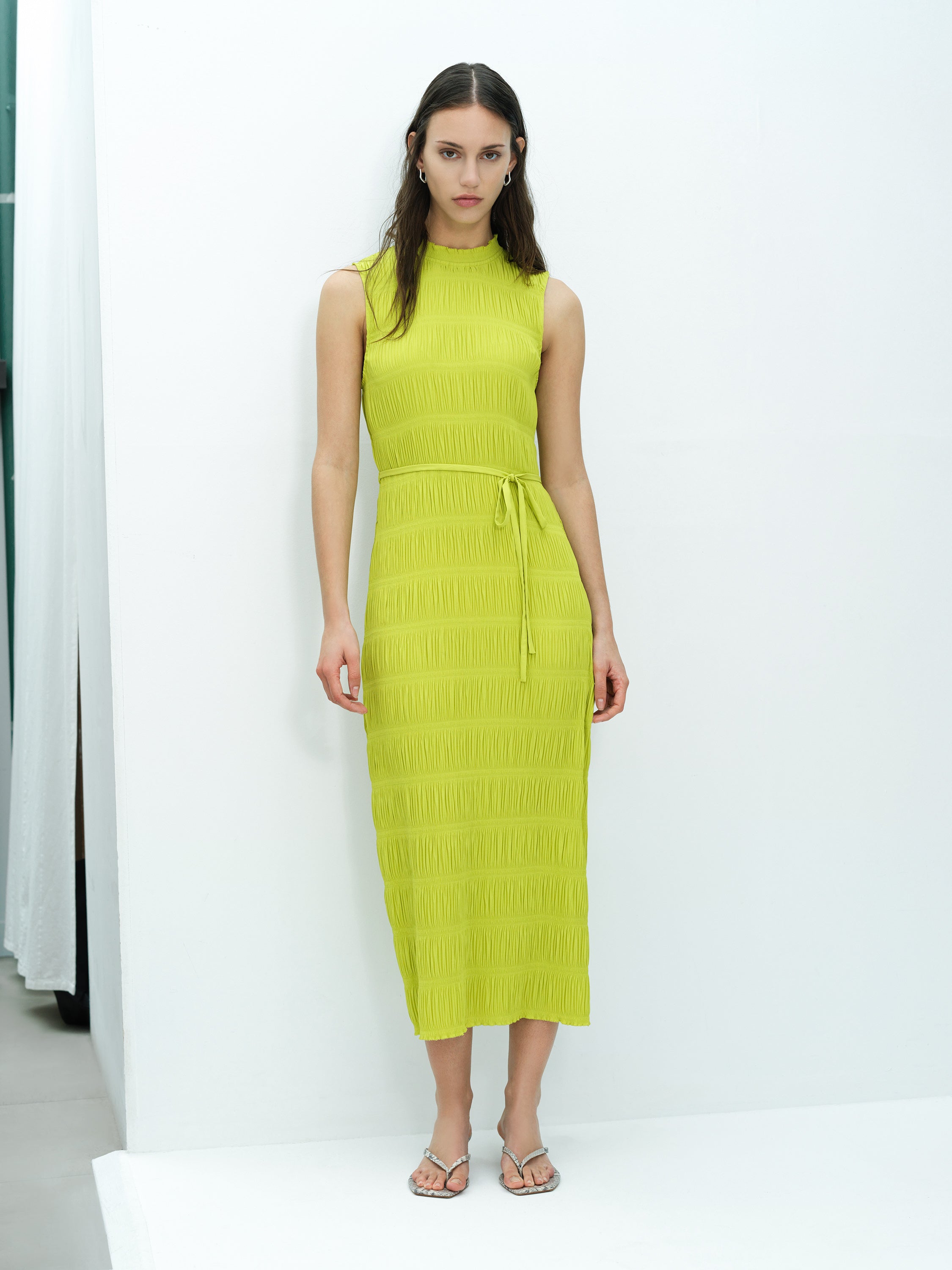A woman wearing a sleeveless green midi length dress with a tie waist, high frill neckline and side split hem.