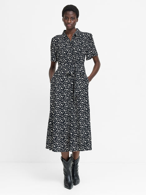 A woman wearing a short sleeved black midi length dress with a shirt collar, button down fastening, self-tie drawstring waist, two pockets and a white floral pattern.