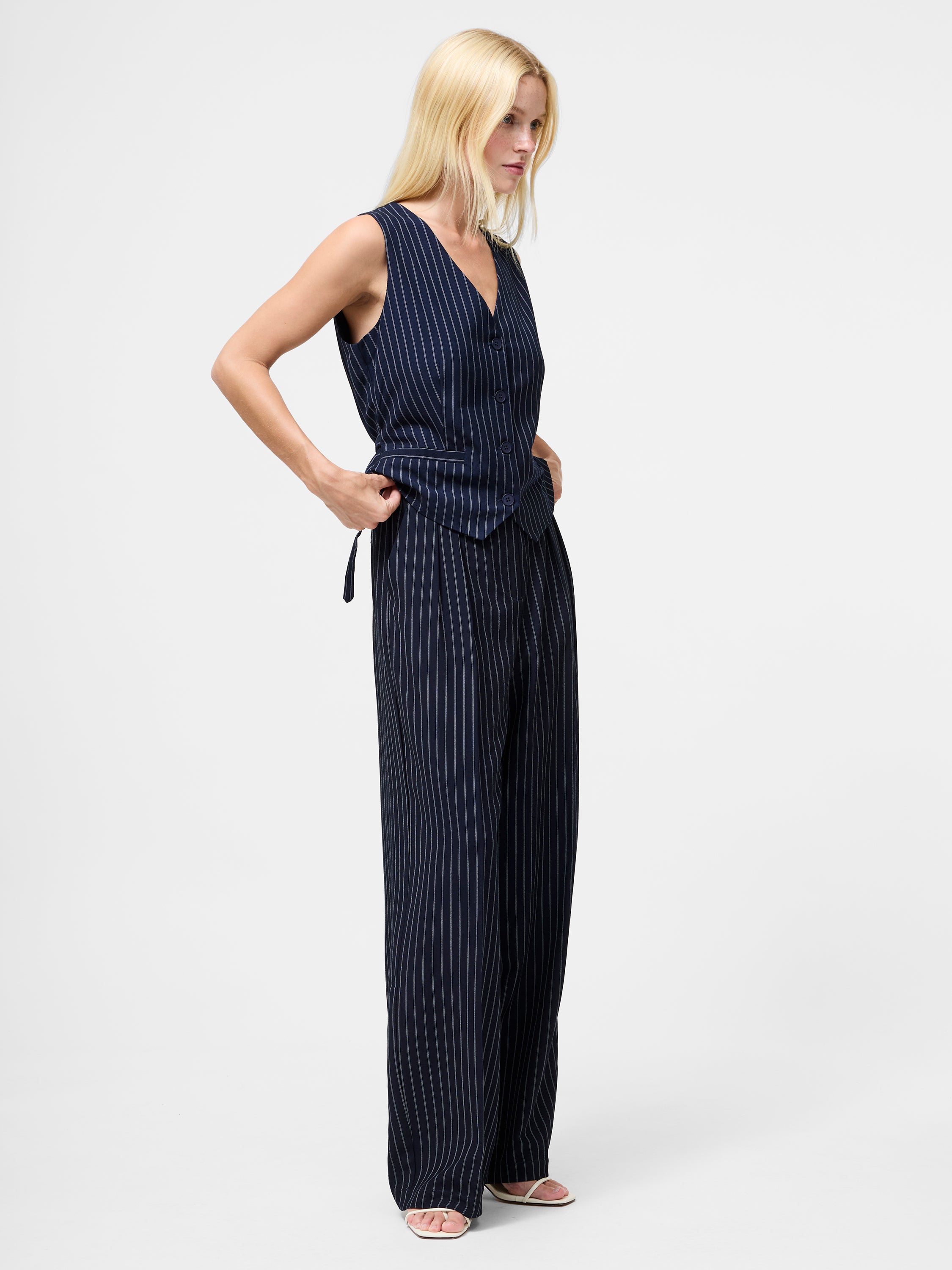 A woman with long blonde hair wears Pinstripe Tailored Wide Leg Trousers and a sleeveless navy vest with open-toe sandals, standing against a plain white background.