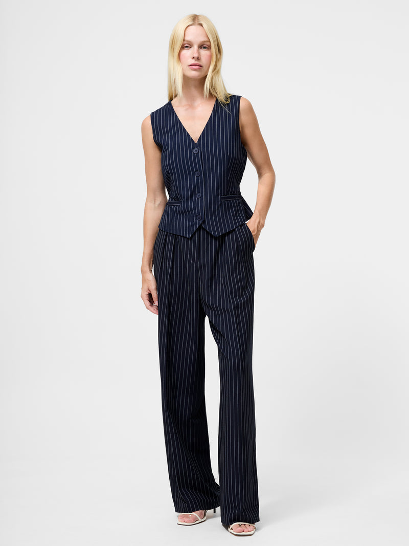 A woman with straight blonde hair stands against a plain background wearing Pinstripe Tailored Wide Leg Trousers, a matching sleeveless vest, and white open-toe heels. Her left hand is in her pocket.