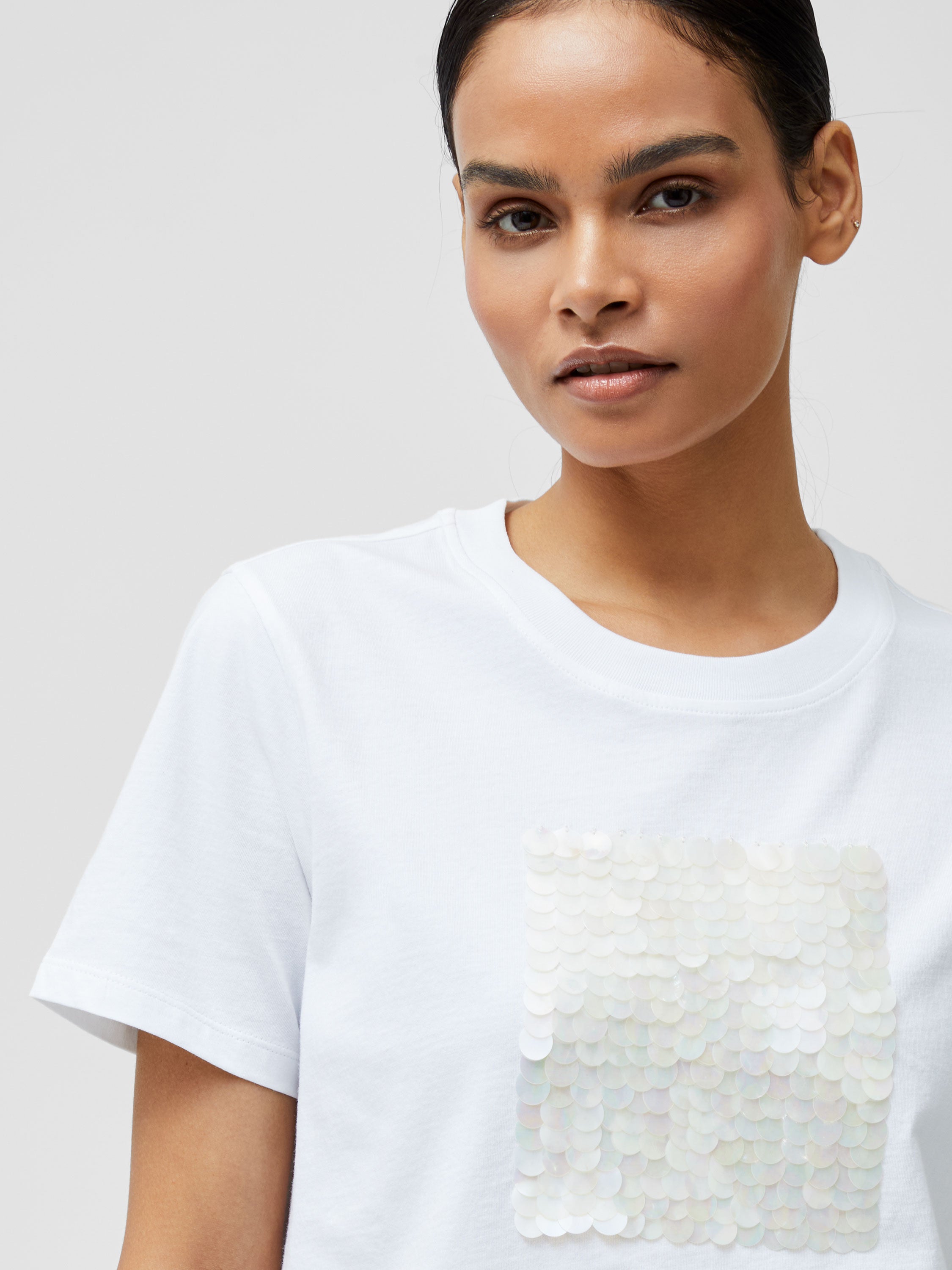 A woman with dark hair pulled back wears the Sequin Jersey Short Sleeve T-Shirt, which features a square sequin design on the chest, standing against a plain light background.