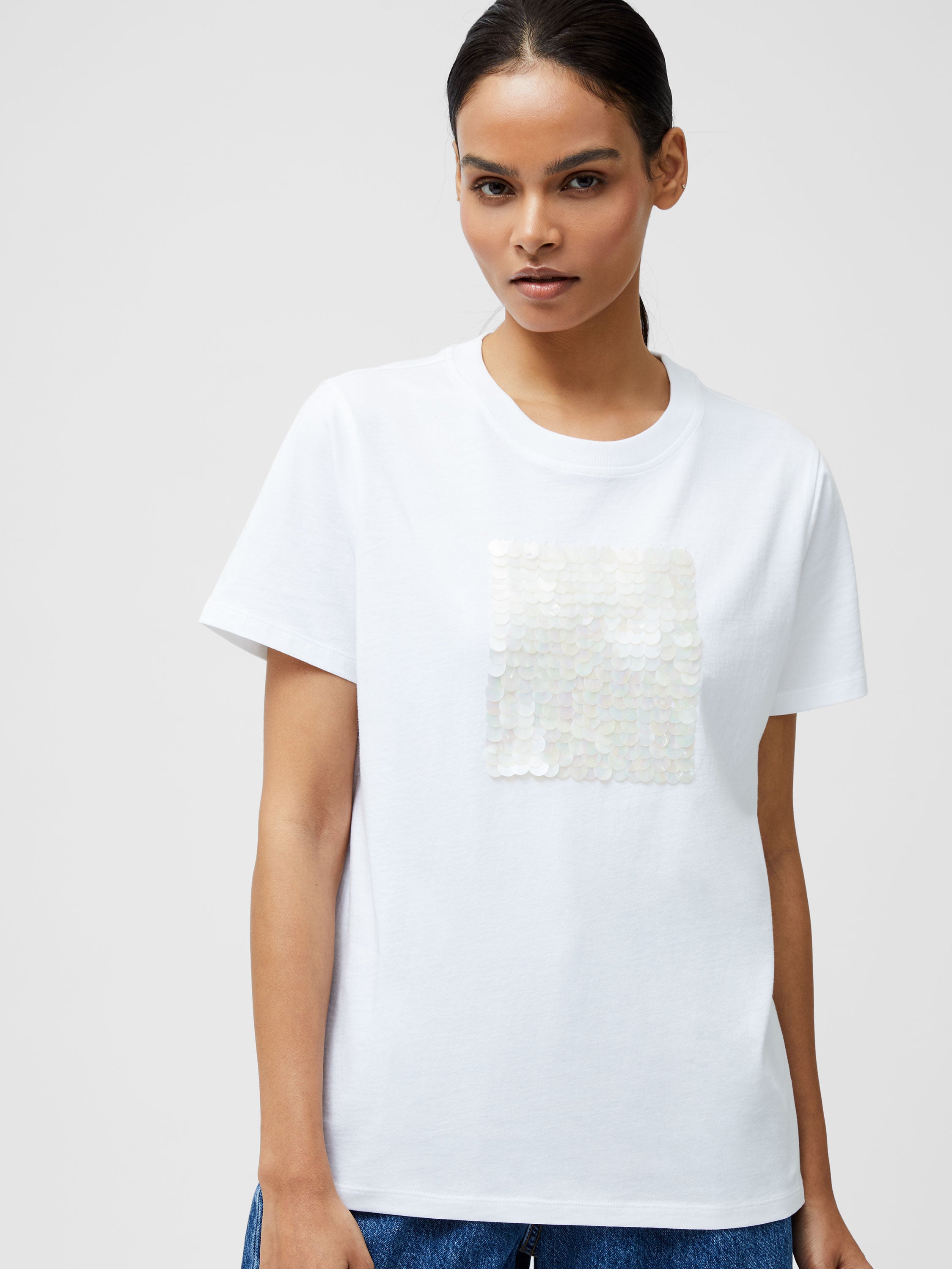 A woman with dark hair pulled back wears the Sequin Jersey Short Sleeve T-Shirt, showcasing a shimmering square sequin front, paired with blue jeans against a plain light background.