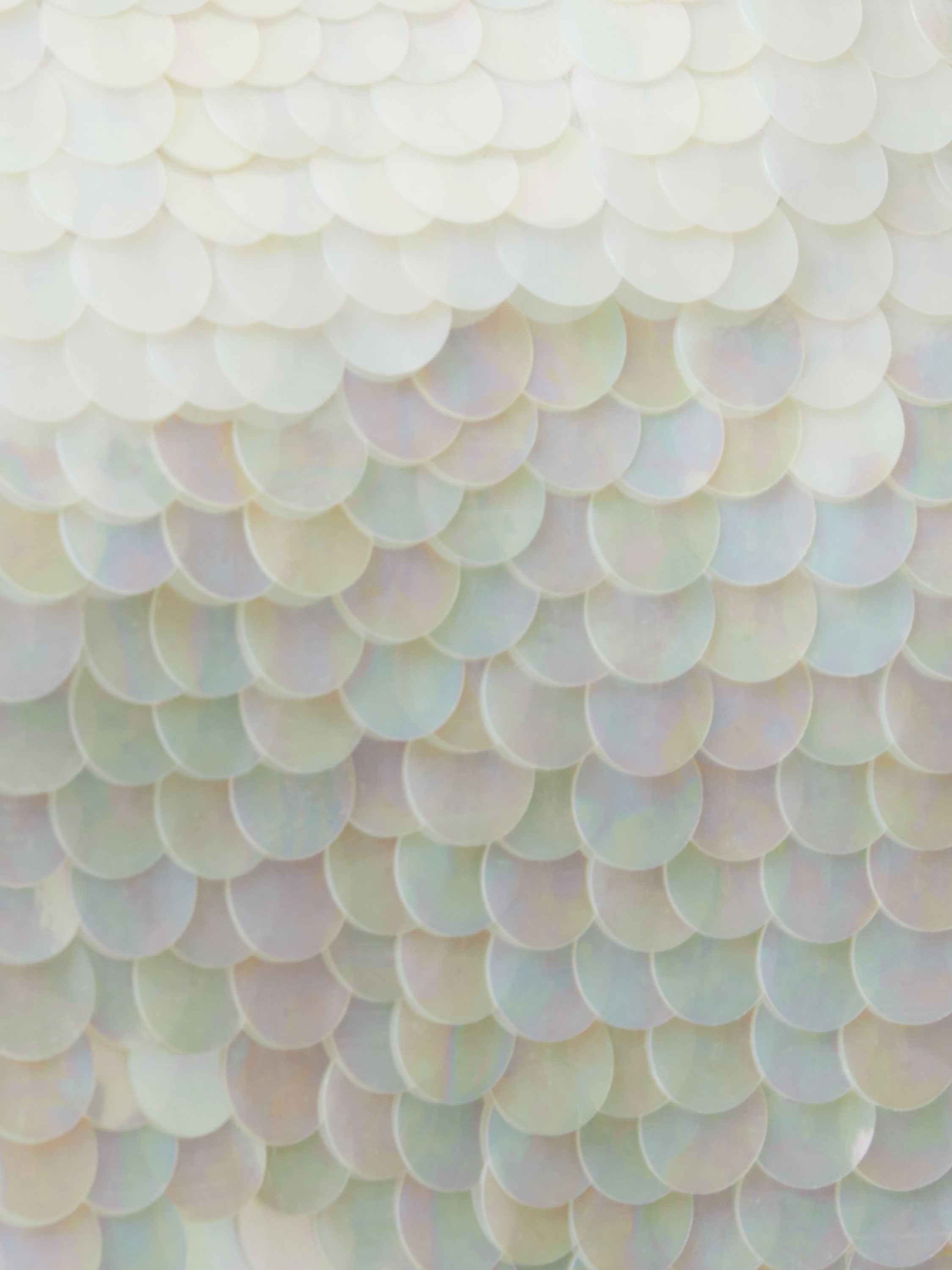 Close-up of overlapping, iridescent sequins in soft pastels on the Sequin Jersey Short Sleeve T-Shirt, creating a shimmering, scale-like texture. Light reflects off square sequins for a pearly, opalescent effect.