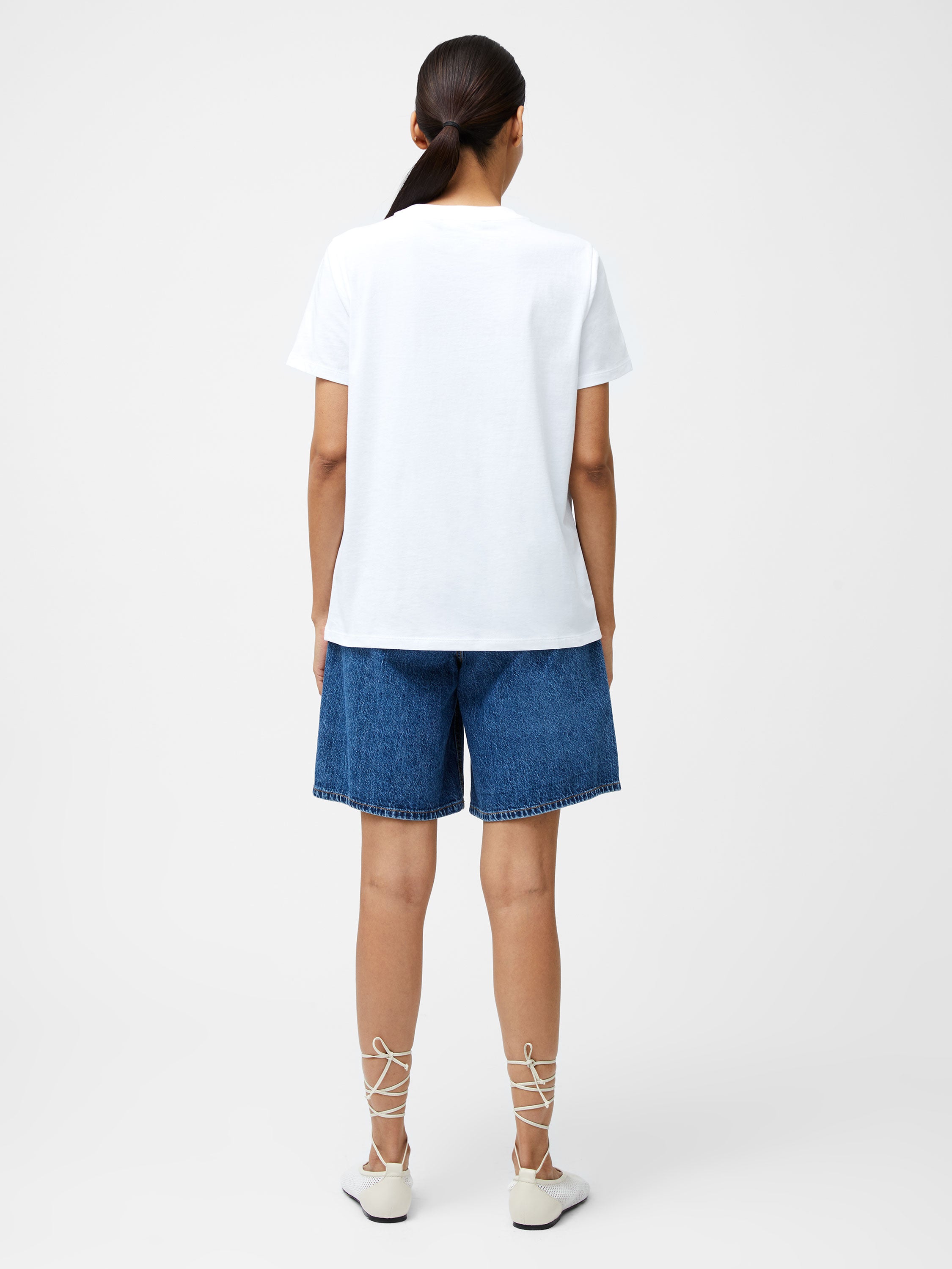 A person stands facing away, wearing a Sequin Jersey Short Sleeve T-Shirt in white, loose blue denim shorts, white sneakers with white lace-up ankle straps, and their hair tied back in a low ponytail.