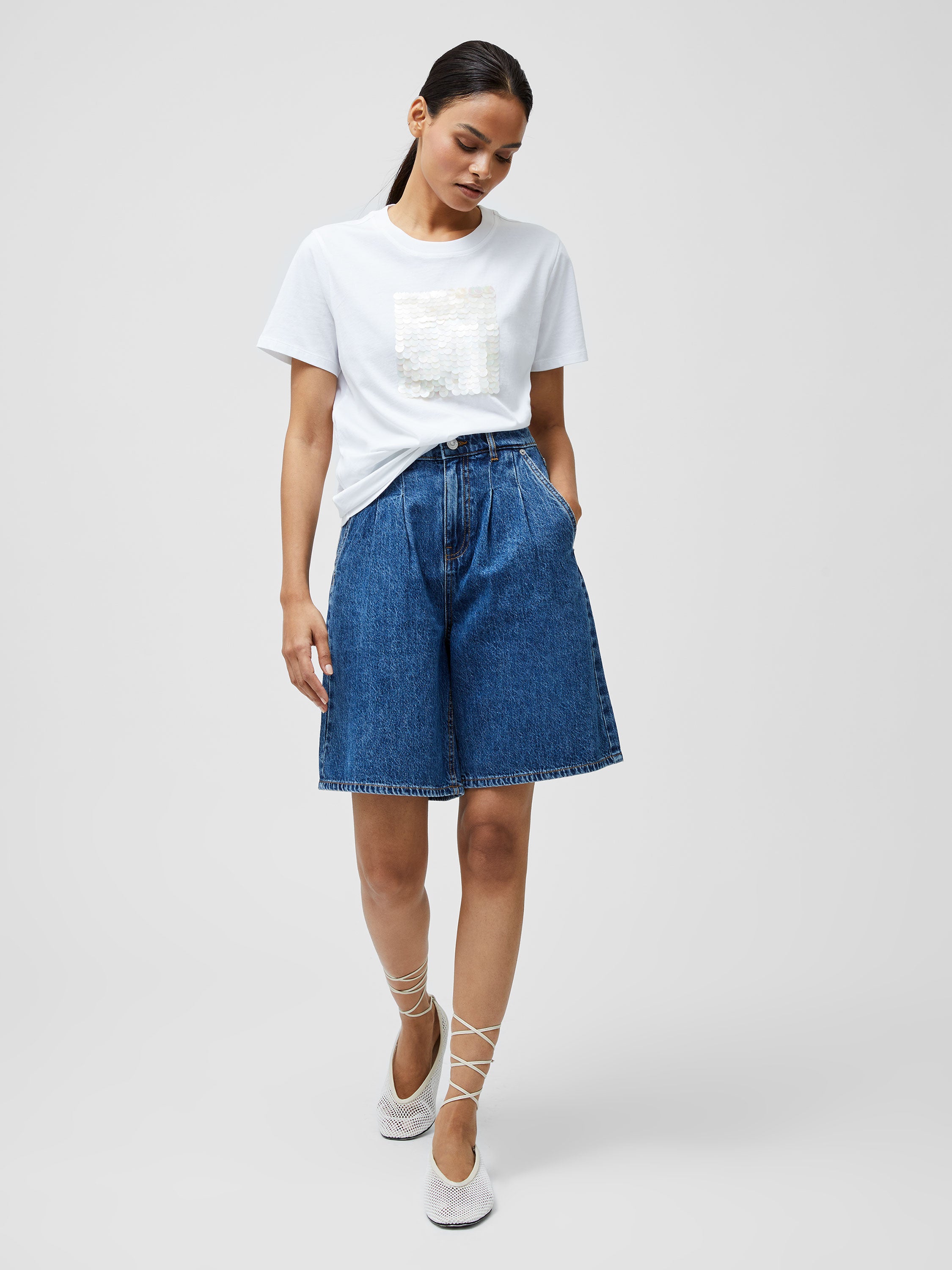A woman with dark hair pulled back stands against a plain background, hands in pockets, wearing a Sequin Jersey Short Sleeve T-Shirt with shiny square sequins, high-waisted blue denim shorts, and white lace-up flats.